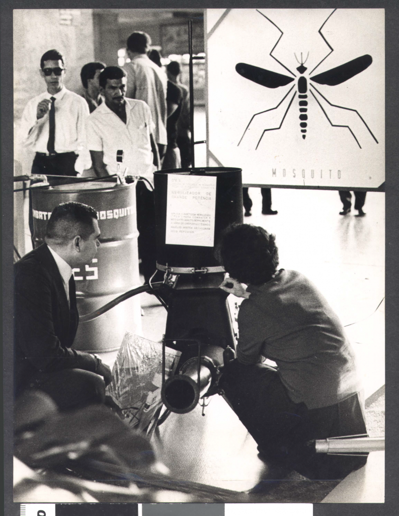 Foto antiga de exposição pública sobre o mosquito, com cartaz e visitantes observando o material