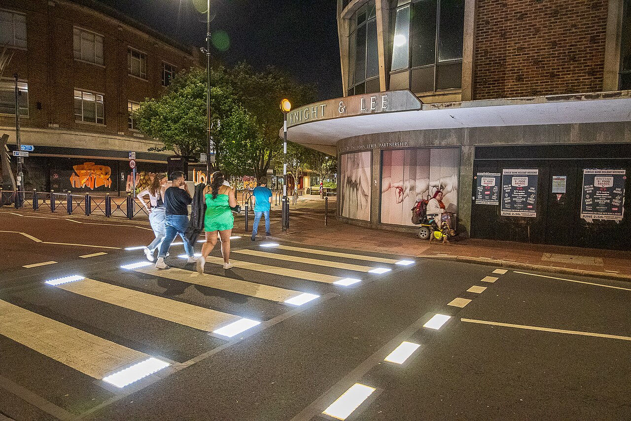 Pedestres atravessando uma rua sobre a faixa de pedestres iluminada, à noite.