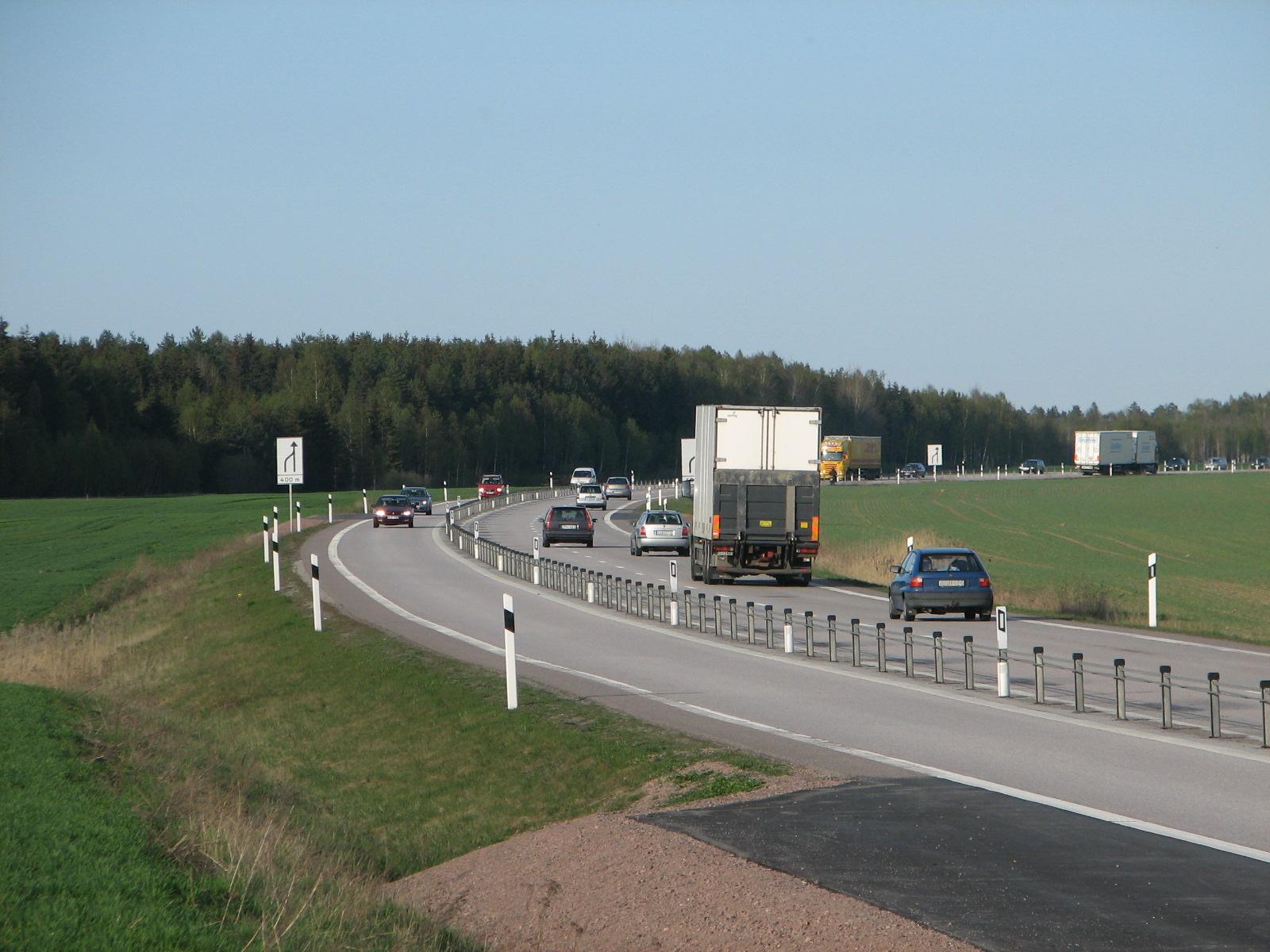Estrada 2+1 sueca (E20) com uma barreira de cabo de aço em postes correndo pelo centro, separando os dois sentidos; caminhões e carros trafegando.