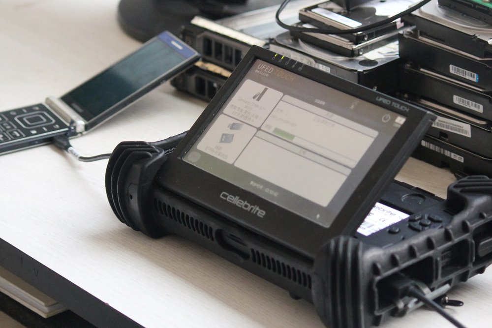 Aparelho Cellebrite UFED Touch sobre uma bancada, com um celular conectado por cabo e discos rígidos ao fundo.